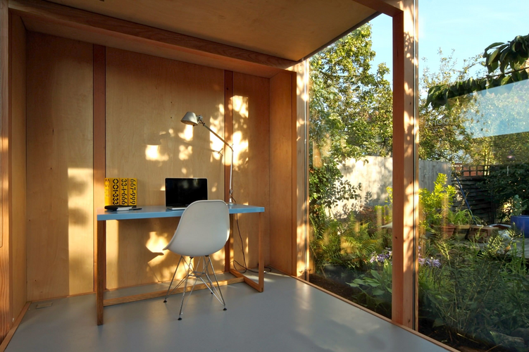 A home office workspace extending onto a shaded balcony through sliding glass doors with weather resistant seating.